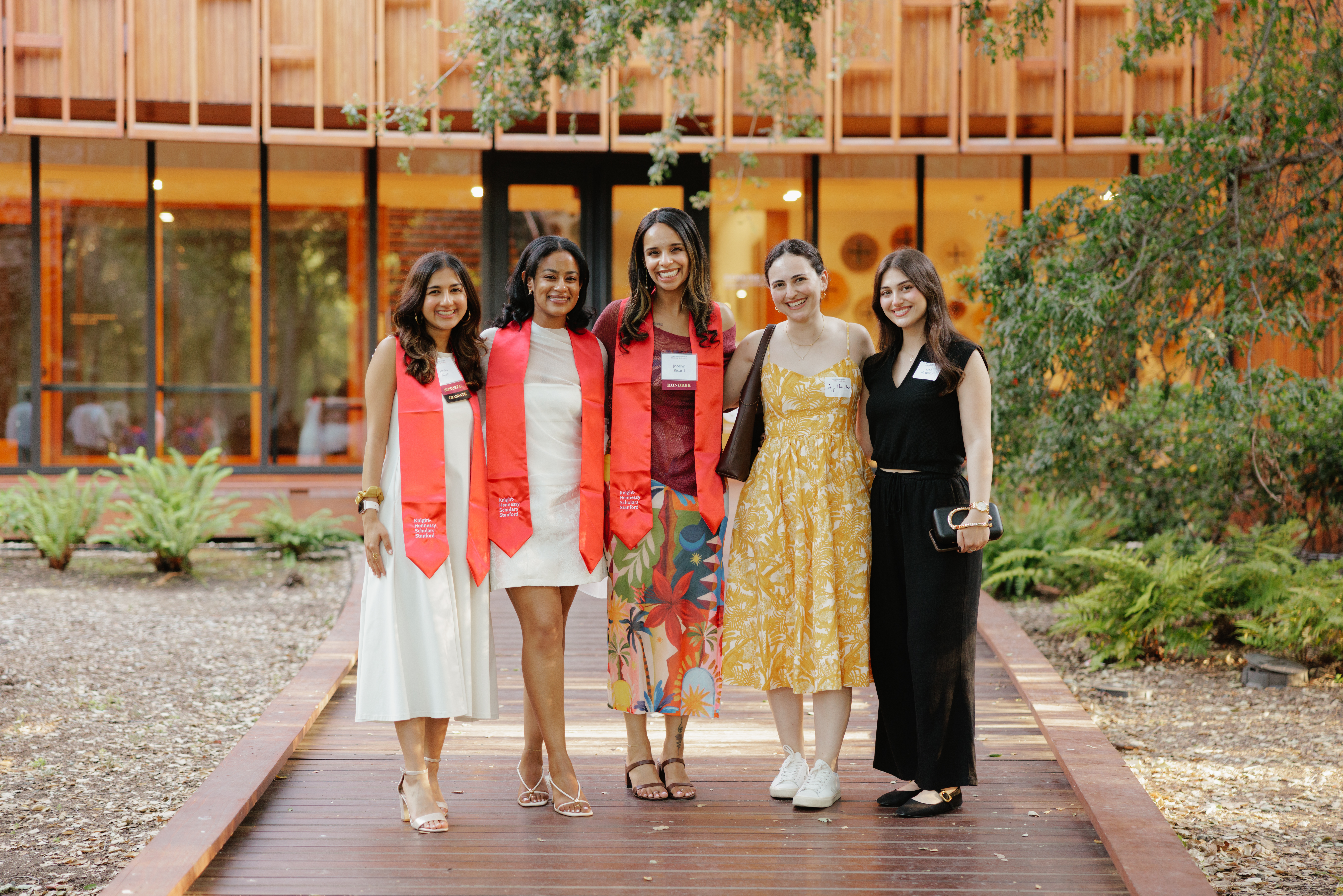 Leona Neftaliem with four fellow Knight-Hennessy Scholars at their Stanford graduation, standing outdoors in front of a wood-clad building surrounded by trees.